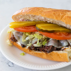 A large sub sandwich has shredded roast beef, melted provolone, shredded lettuce, sliced tomatoes, and thin sliced pickles stacked for the filings.
