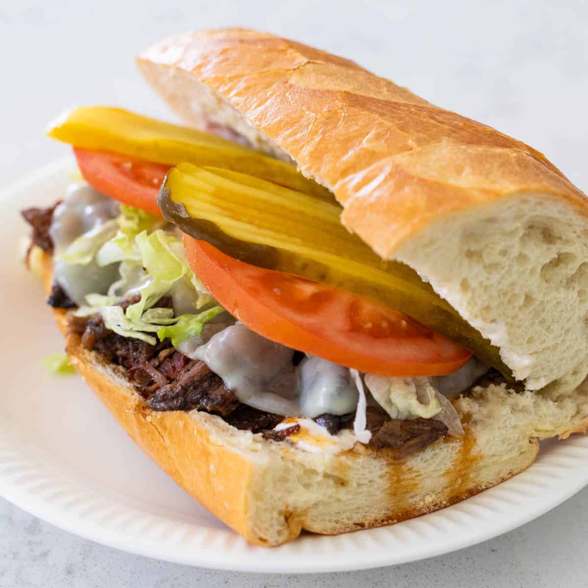 A large sub sandwich has shredded roast beef, melted provolone, shredded lettuce, sliced tomatoes, and thin sliced pickles stacked for the filings.