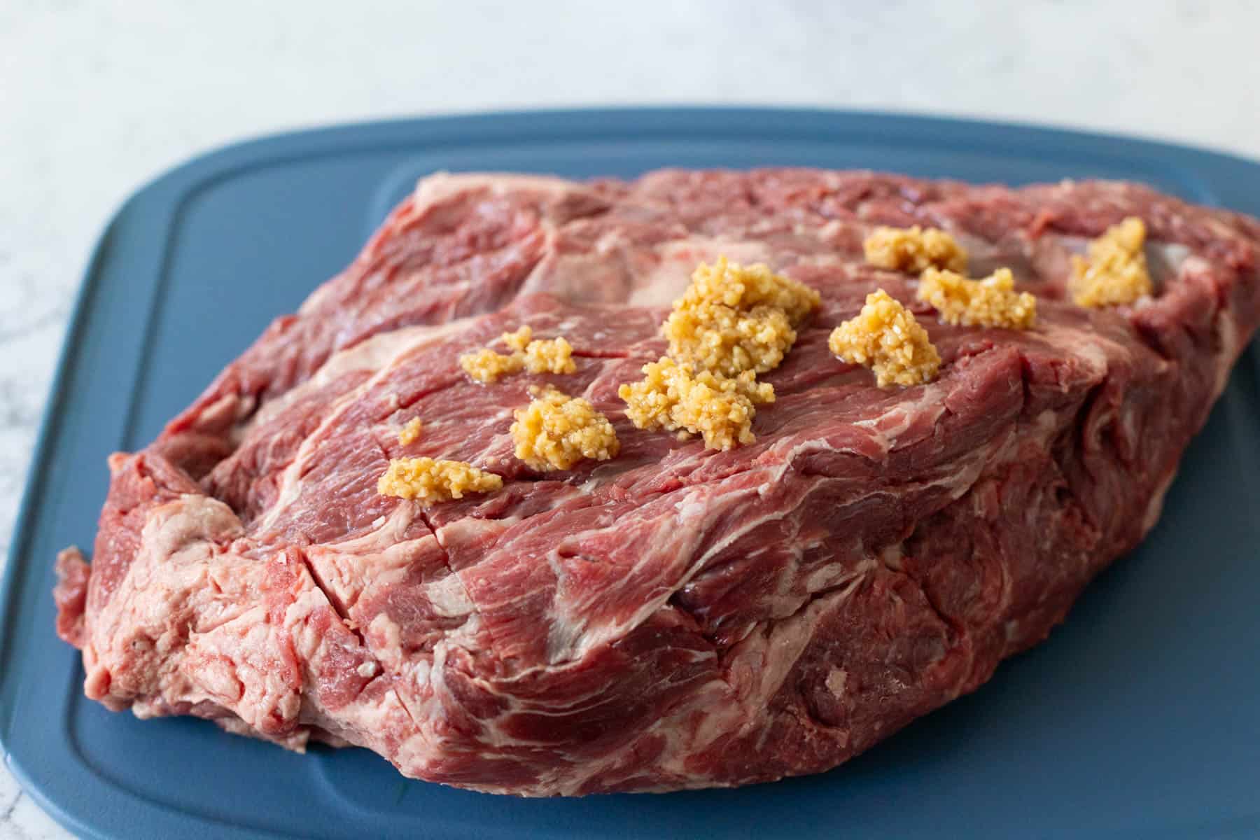 The chuck roast has had shallow cuts all over the surface and the garlic is on top about to be rubbed in.