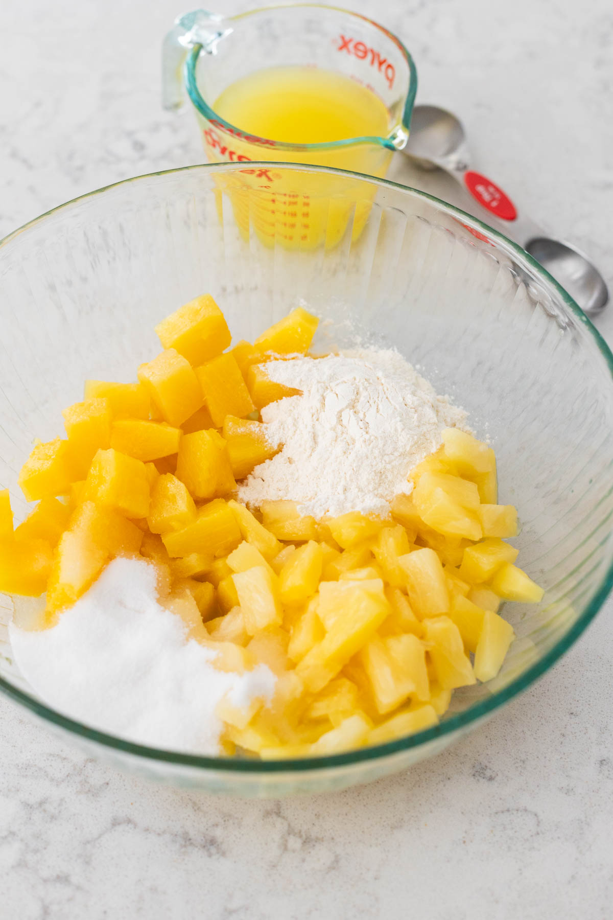 The mixing bowl has pineapple, sugar, and flour. A measuring cup of pineapple juice is in the back.