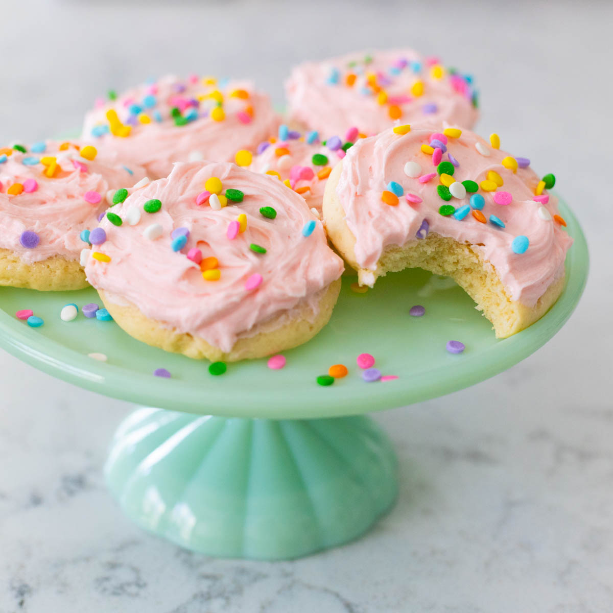 An aqua cake plate has 5 soft frosted sugar cookies decorated with pink icing and sprinkles. One cookie has a bite taken out of it.