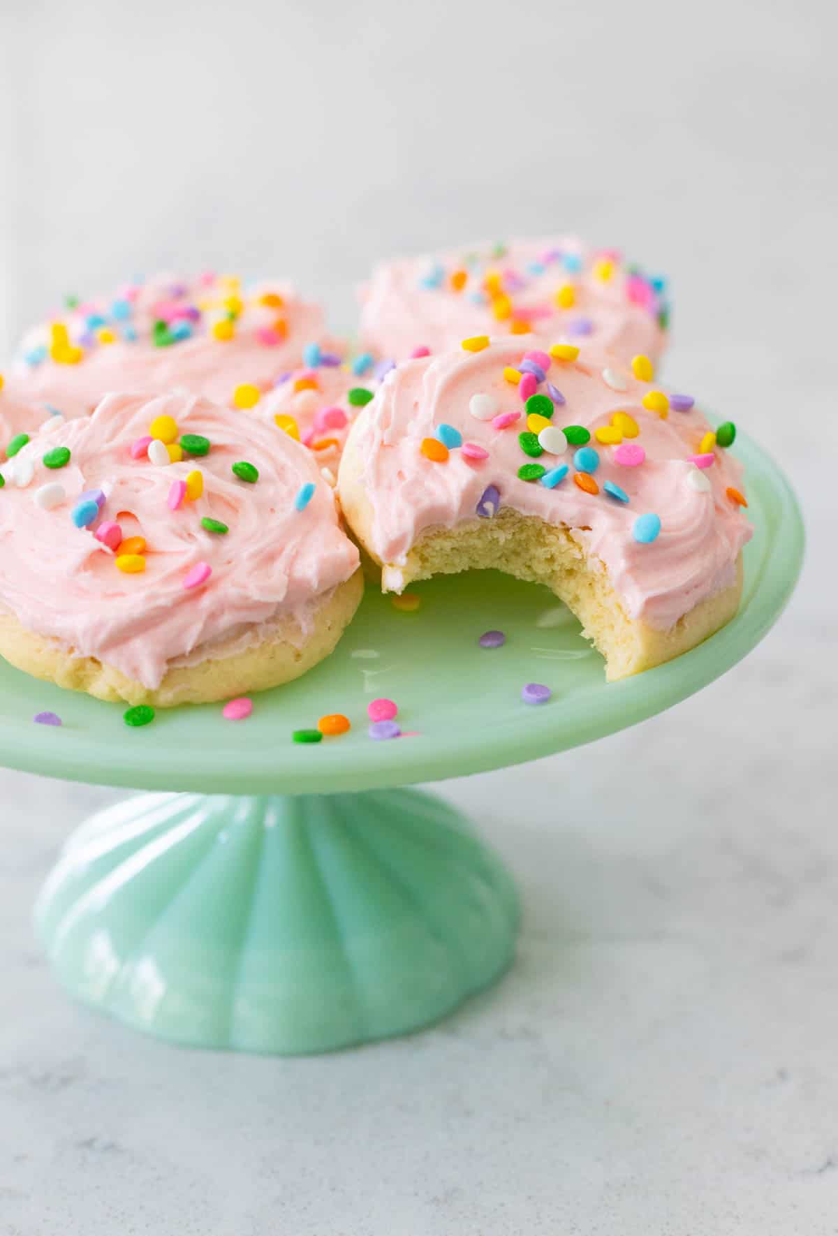 An aqua cake plate has 5 soft frosted sugar cookies decorated with pink icing and sprinkles. One cookie has a bite taken out of it.