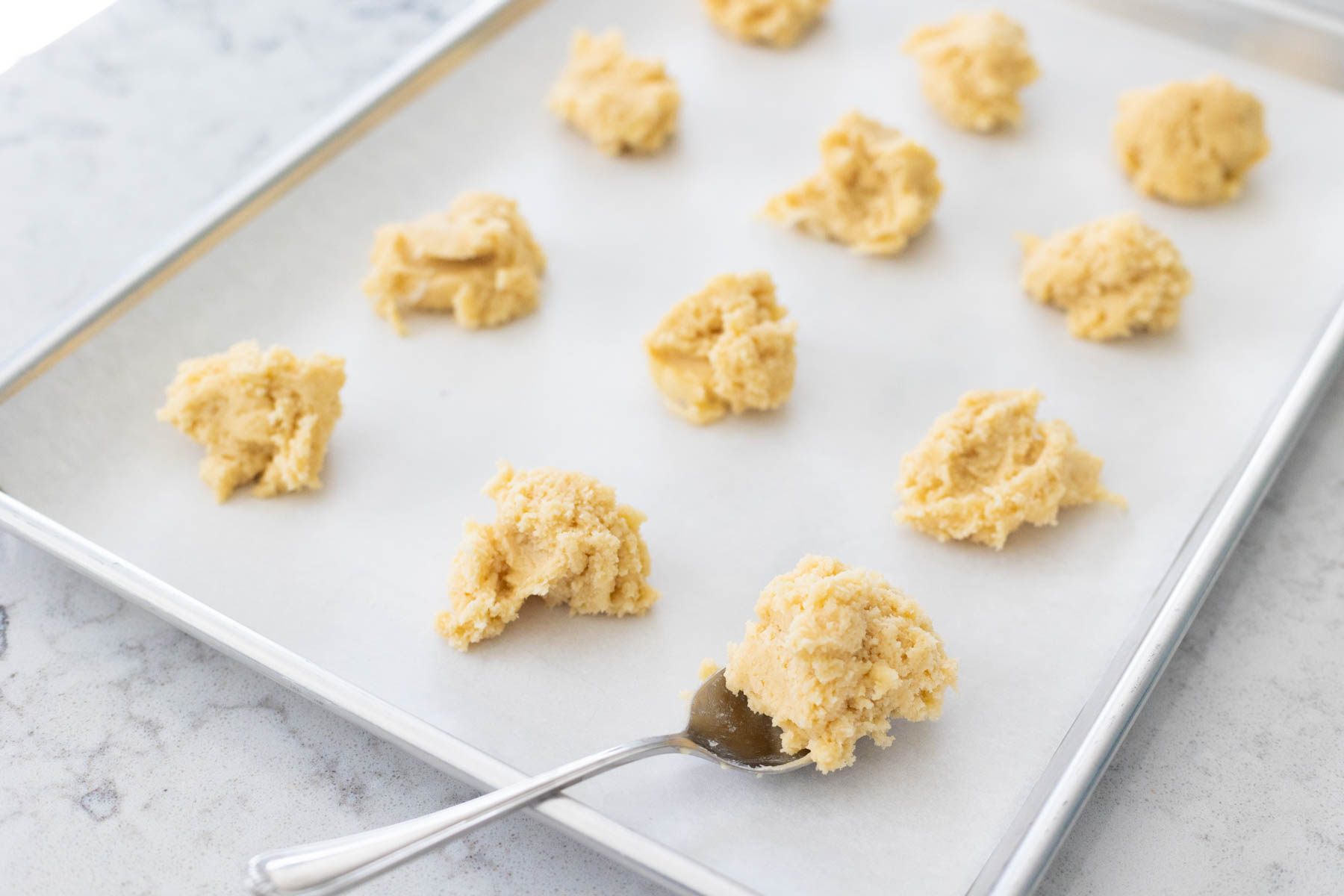 A baking sheet lined with parchment paper has a dozen sugar cookie balls in place. A regular table spoon is portioning out a ball of dough in the corner.