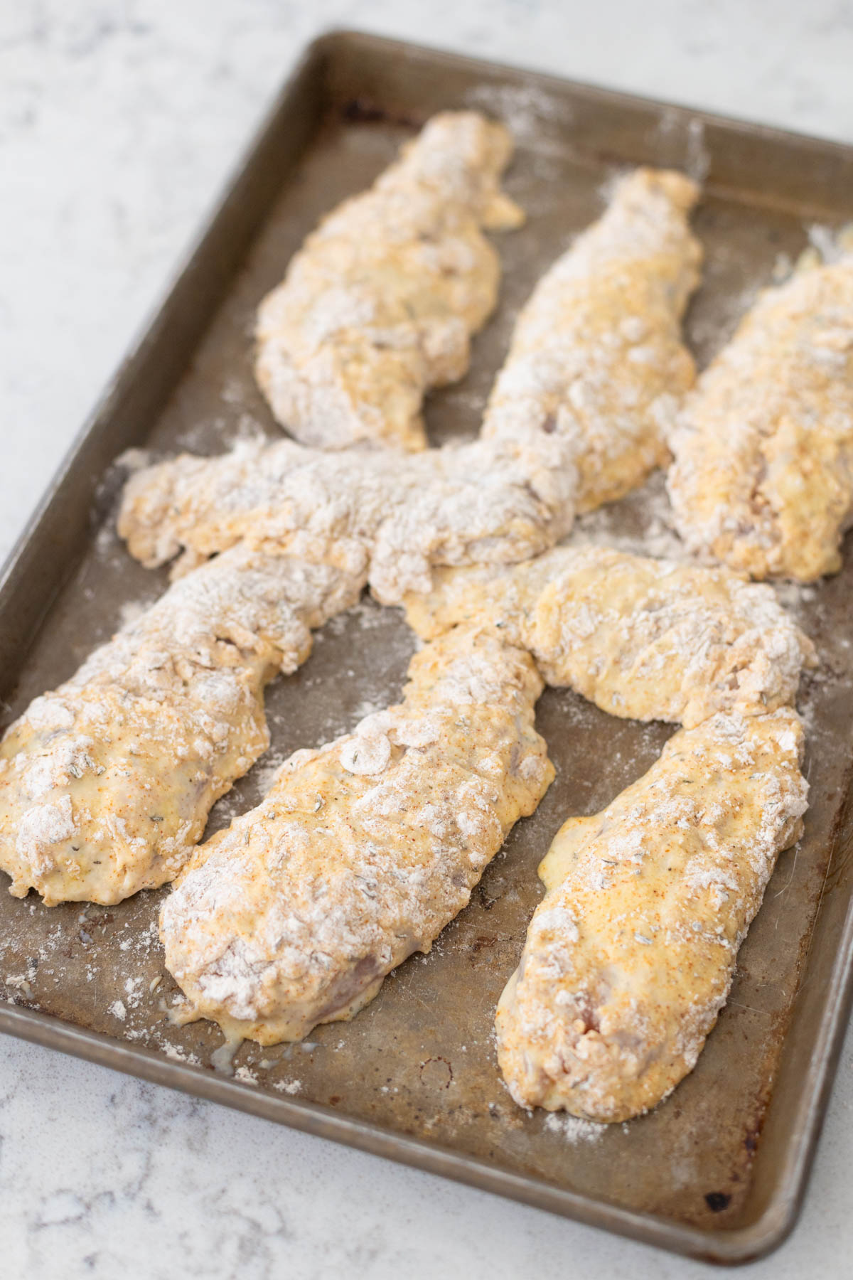 The chicken tenders have been breaded and are lined up on a baking pan.