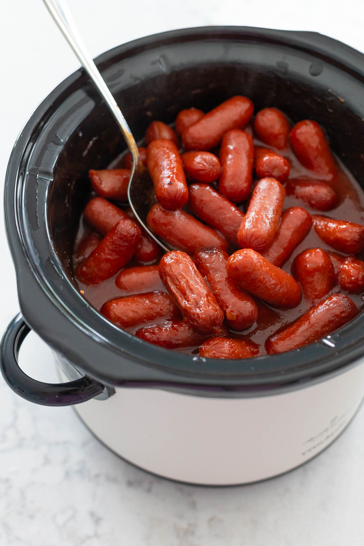 Saucy little hot dogs are in a slowcooker being stirred by a spoon.