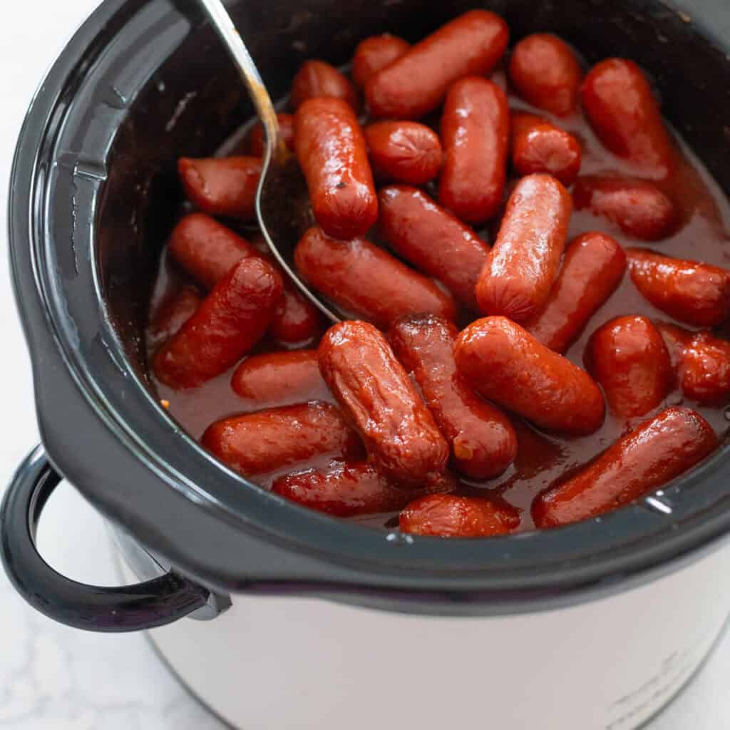 Saucy little hot dogs are in a slowcooker being stirred by a spoon.