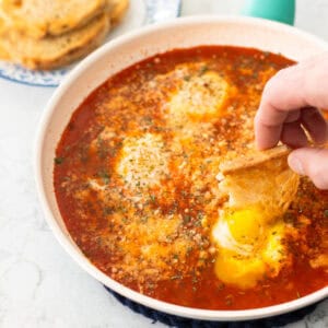 A piece of toast is being dunked into a soft egg in a rich tomato sauce.