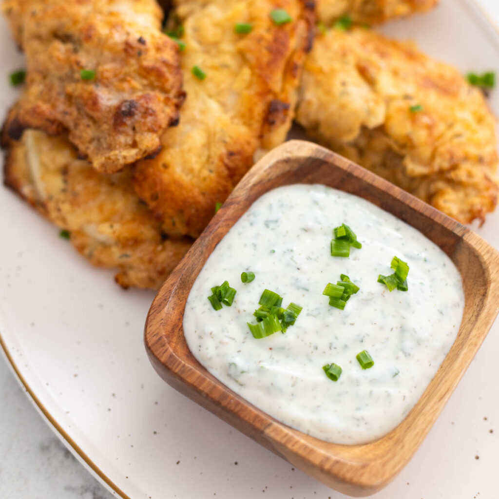 A bowl of ranch dip with fresh chives on top sits next to a pile of fried chicken.