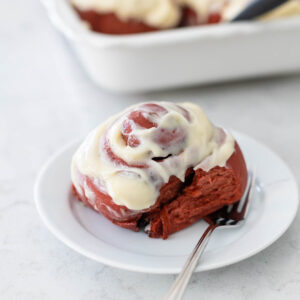 A red velvet cinnamon roll with cream cheese frosting is on a white plate with a fork.