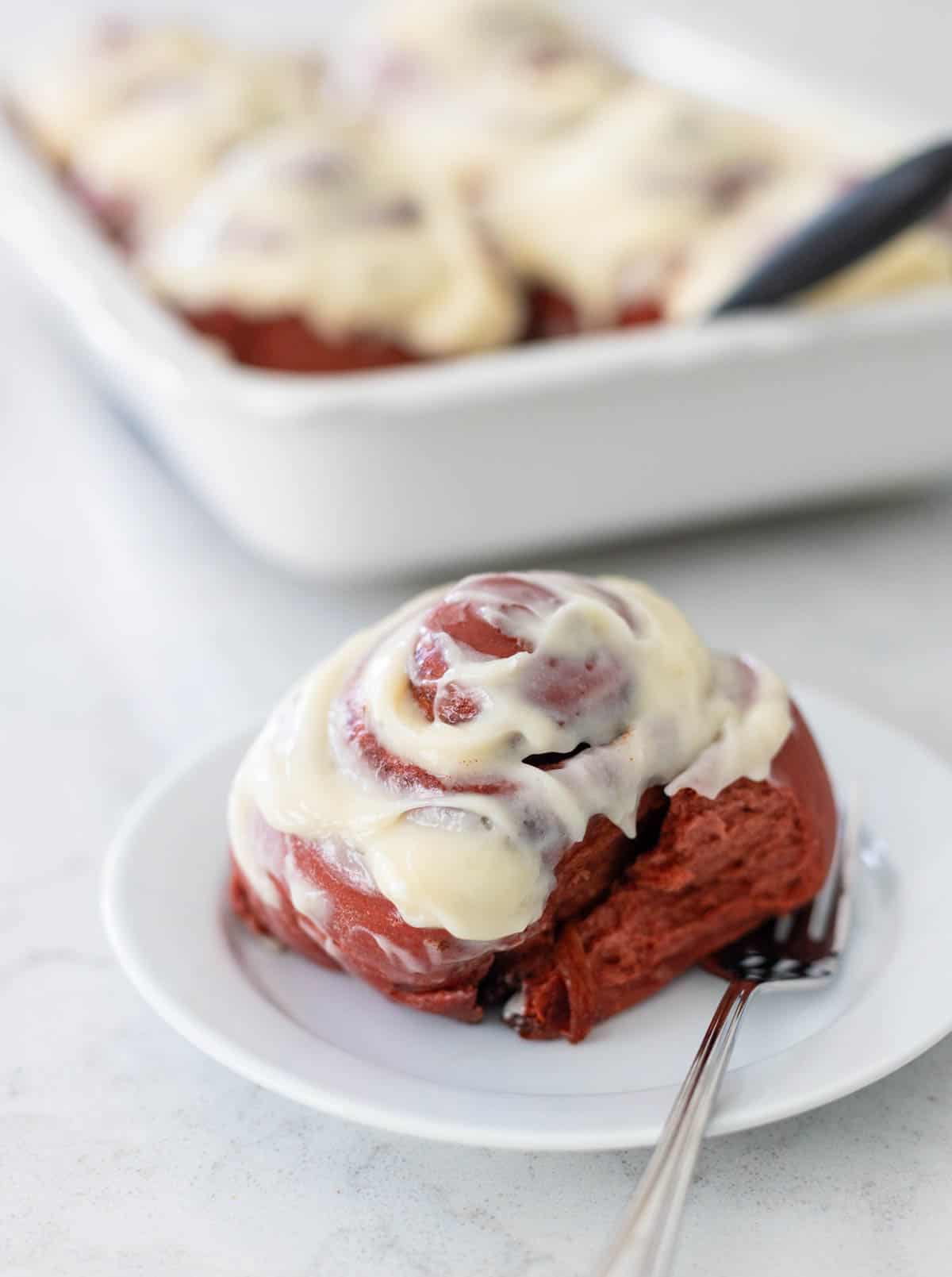 A singled cinnamon roll is on a white plate with a fork next to the baking pan filled with rolls.