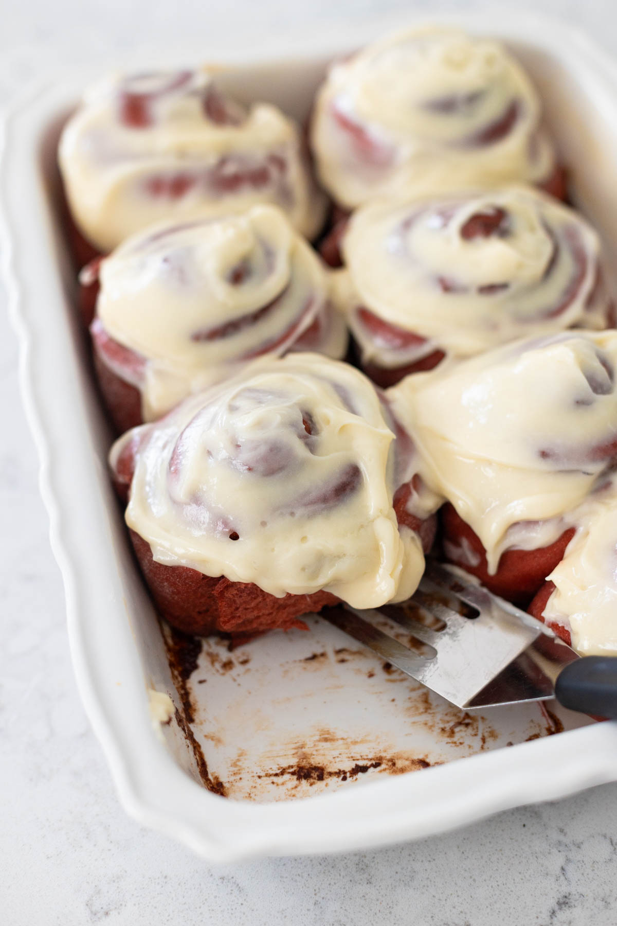 A pan of red velvet cinnamon rolls with cream cheese icing has one bun missing.