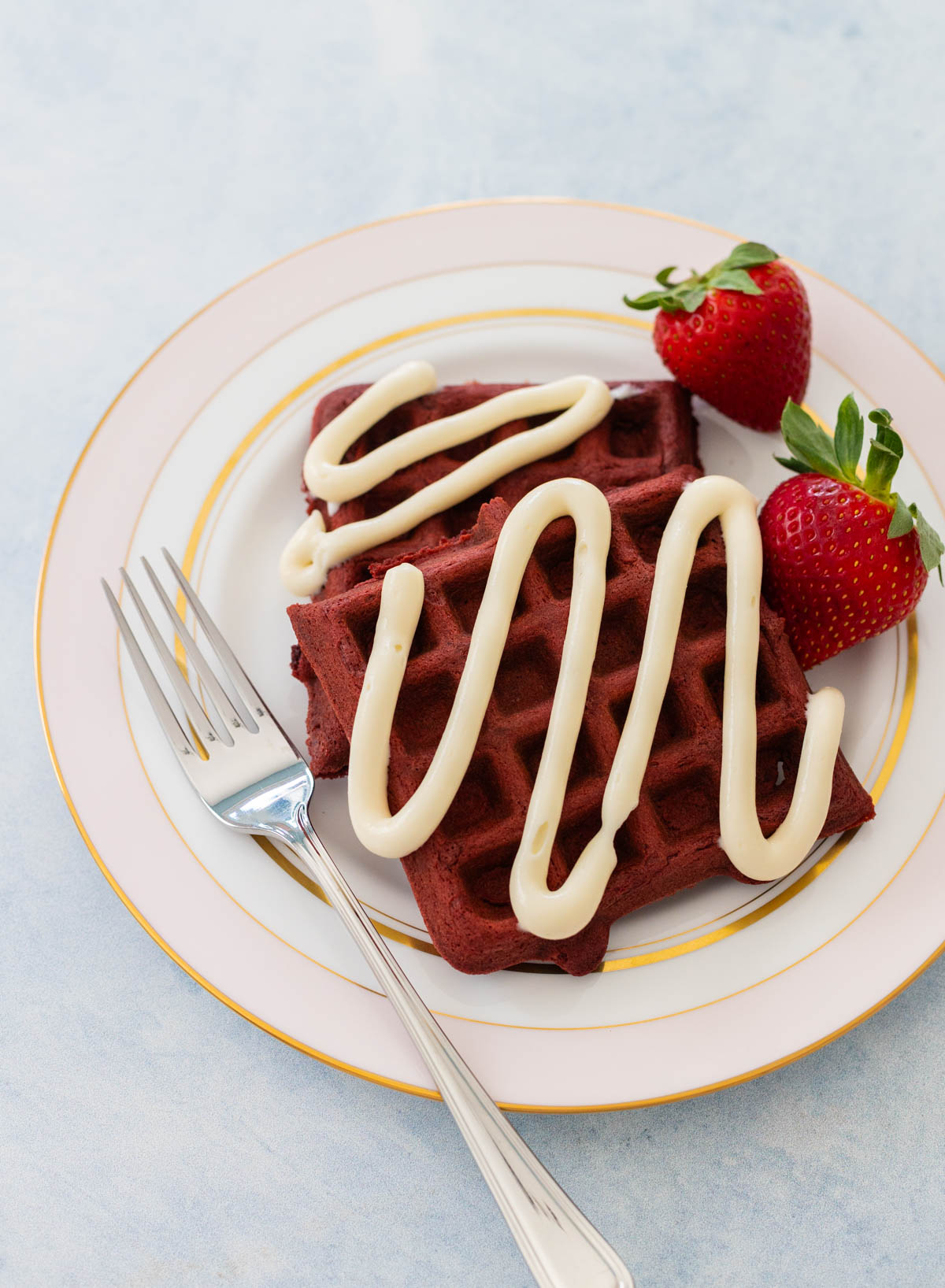 A plate of red velvet waffles has a drizzle of cream cheese icing and fresh strawberries on the side.