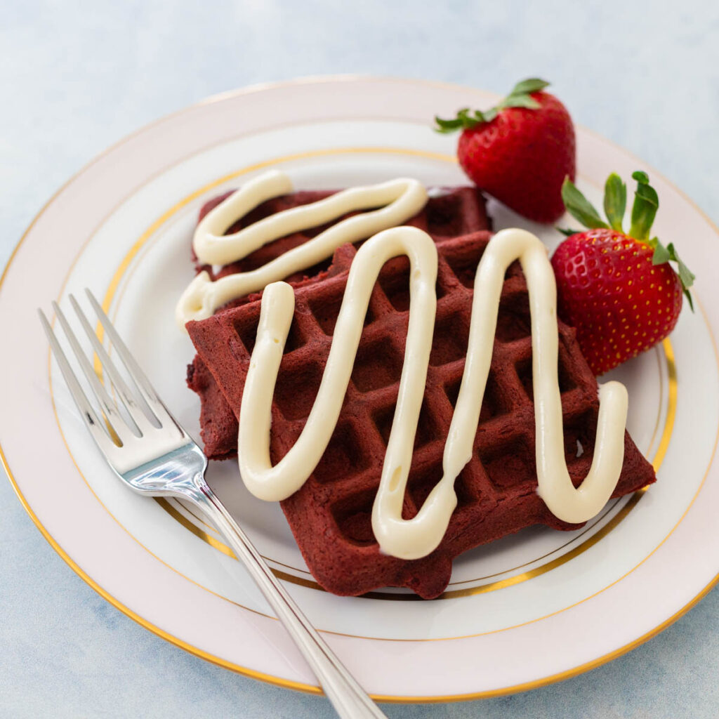 A plate of red velvet waffles has a drizzle of cream cheese icing and fresh strawberries on the side.