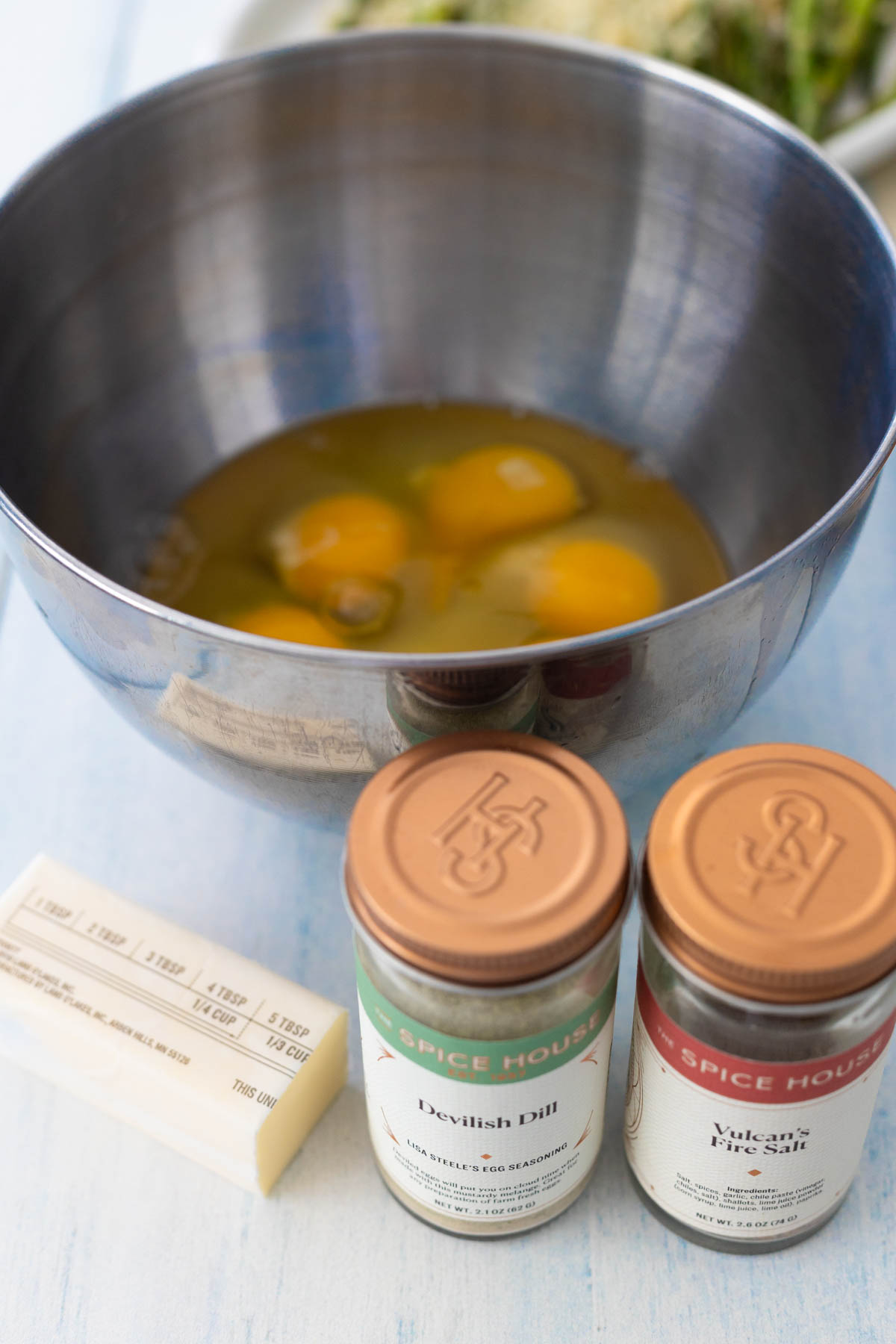The eggs have been cracked into a mixing bowl that sits next to a stick of butter and two jars of spices.
