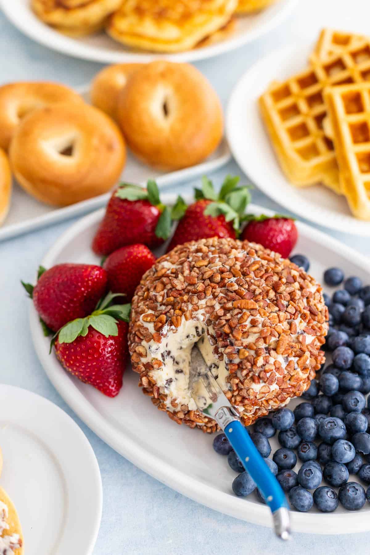 A cream cheese and chocolate chip cheese ball coated in pecans sits next to fresh fruit and a bagel.