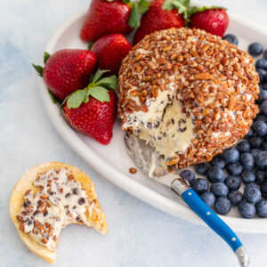 A cream cheese and chocolate chip cheese ball coated in pecans sits next to fresh fruit and a bagel.