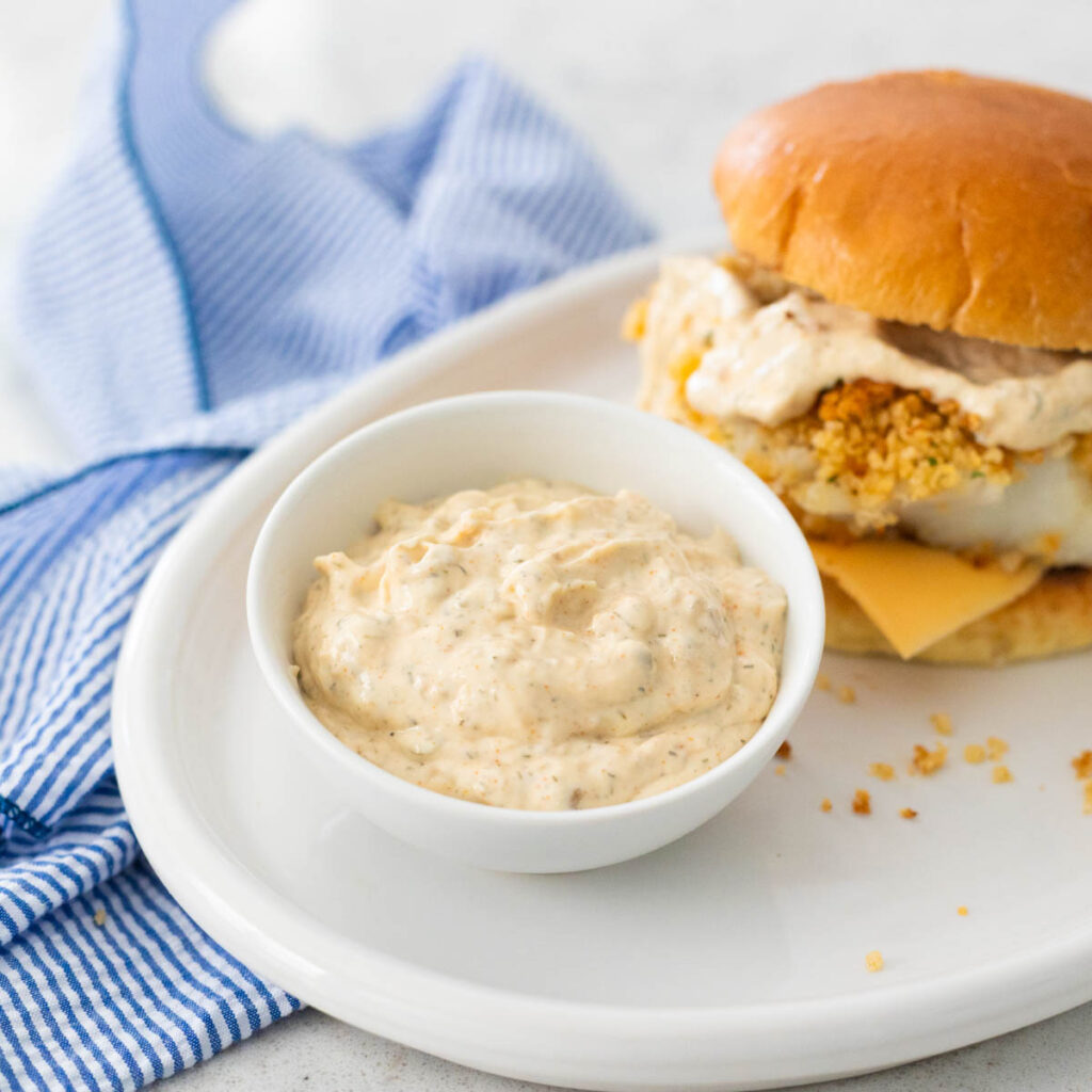 The cup of tartar sauce is on a platter next to a fish sandwich.