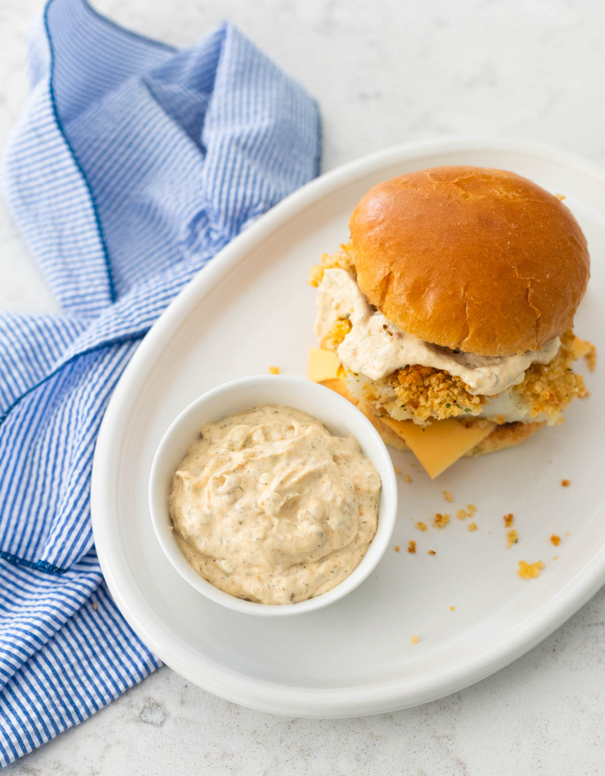 The tartar sauce is in a small cup sitting on a plate next to a fish sandwich.