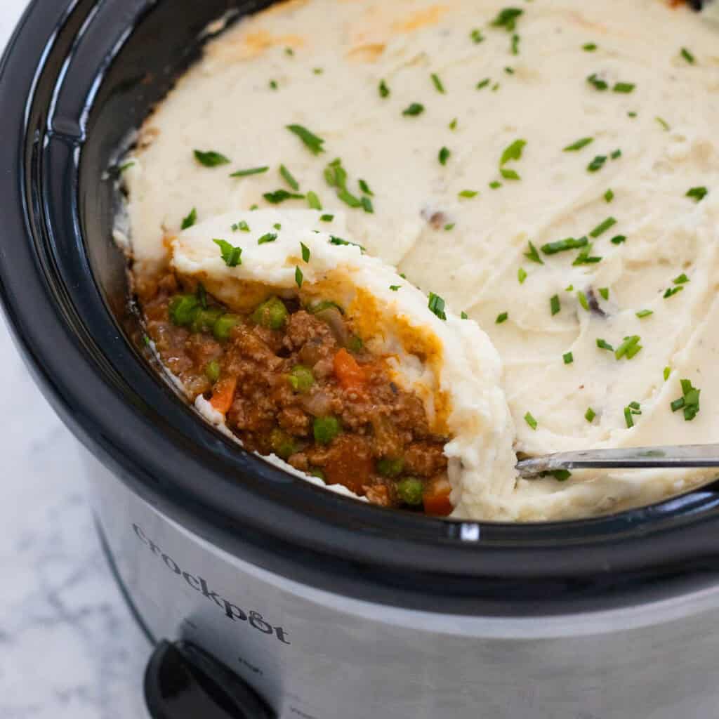 A slowcooker is filled with a beef stew topped with mashed potatoes and parsley.
