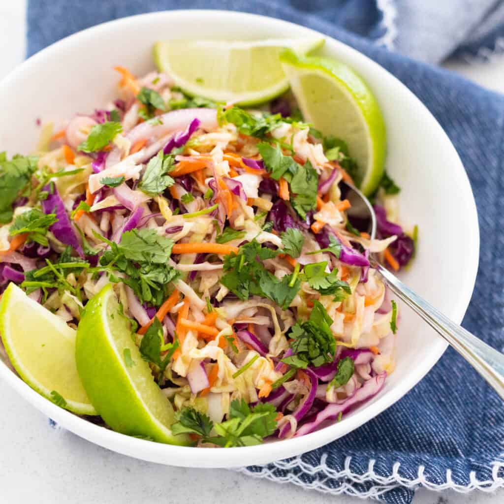 The slaw is in a white bowl with a spoon. Fresh lime wedges are on the side.