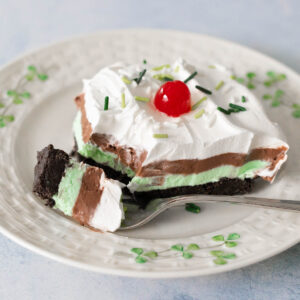 A layered dessert with oreo cookie crust, creamy green layer, chocolate pudding layer, and whipped topping with a cherry on top on a shamrock plate.