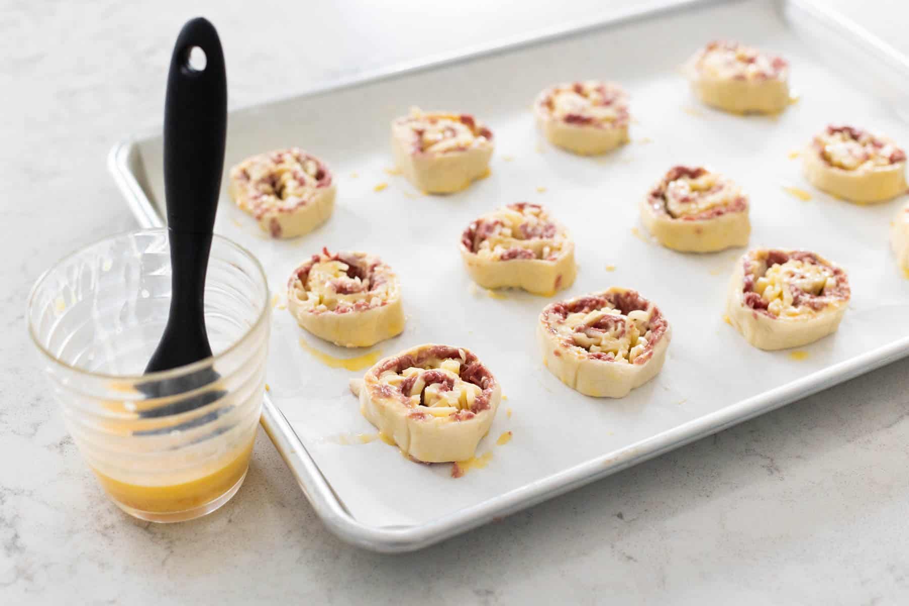 A baking sheet lined with parchment has cut slices of the reuben log and a cup of egg wash on the side with a pastry brush.