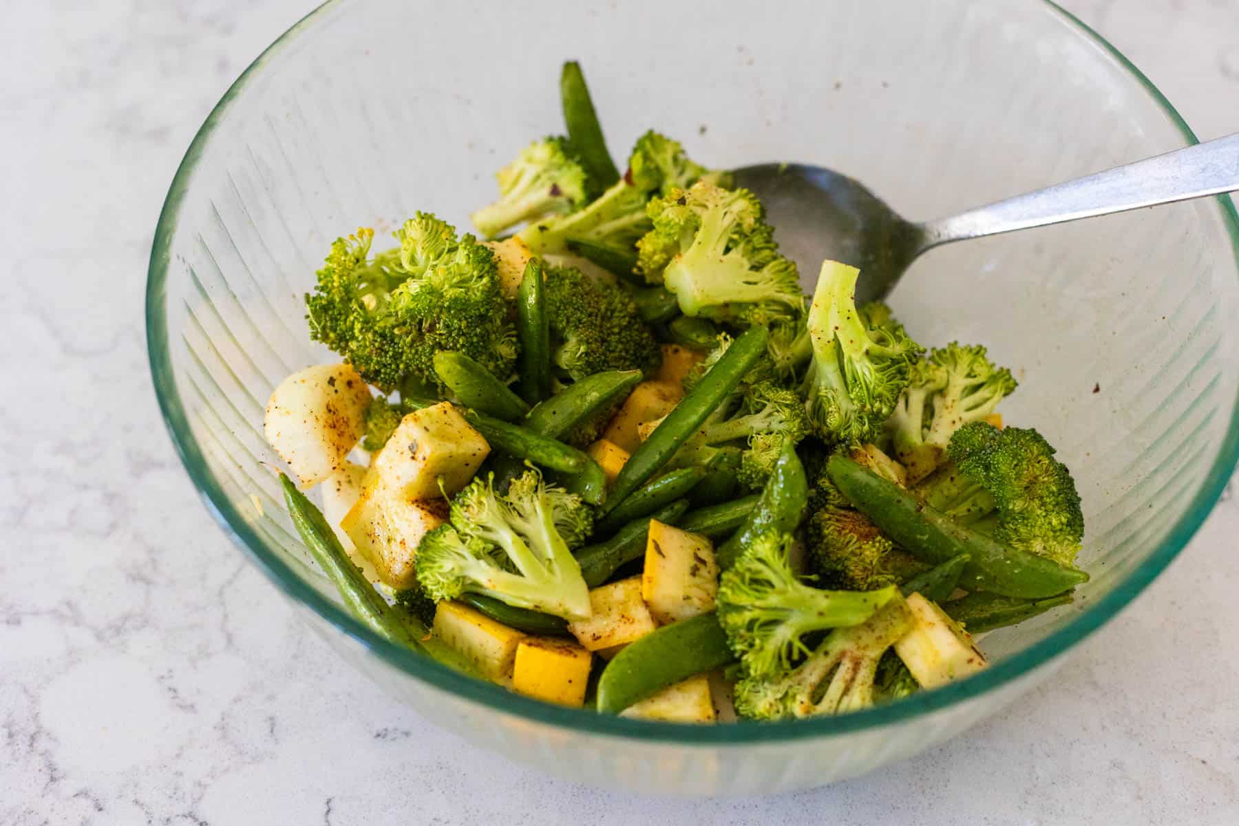 A mixing bowl has all the veggies tossed with olive oil and seasonings, a spoon is mixing them together.