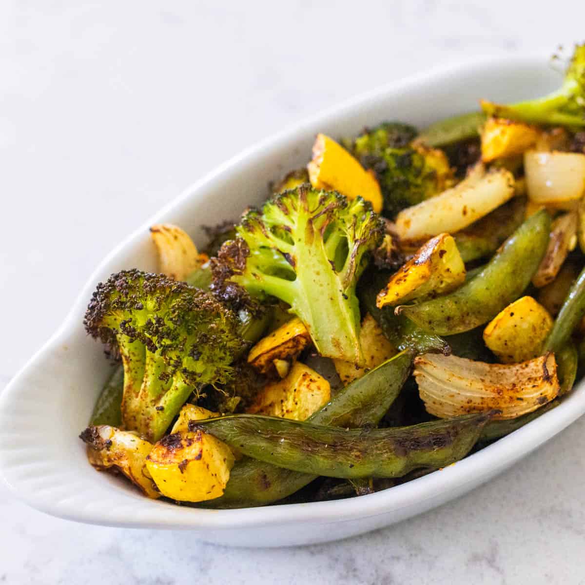 A white dish is filled with broccoli, yellow squash, peas, and onions.