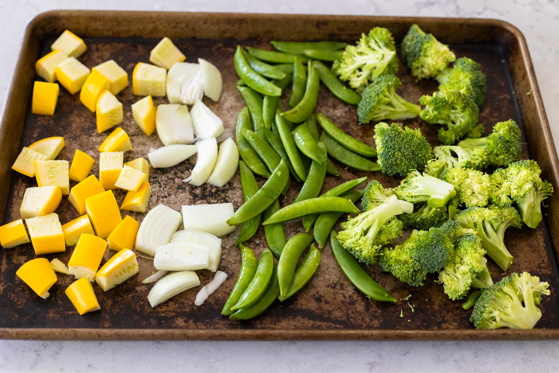 A sheet pan has yellow squash, onion, green peas, and broccoli florets arranged in neat rows. 