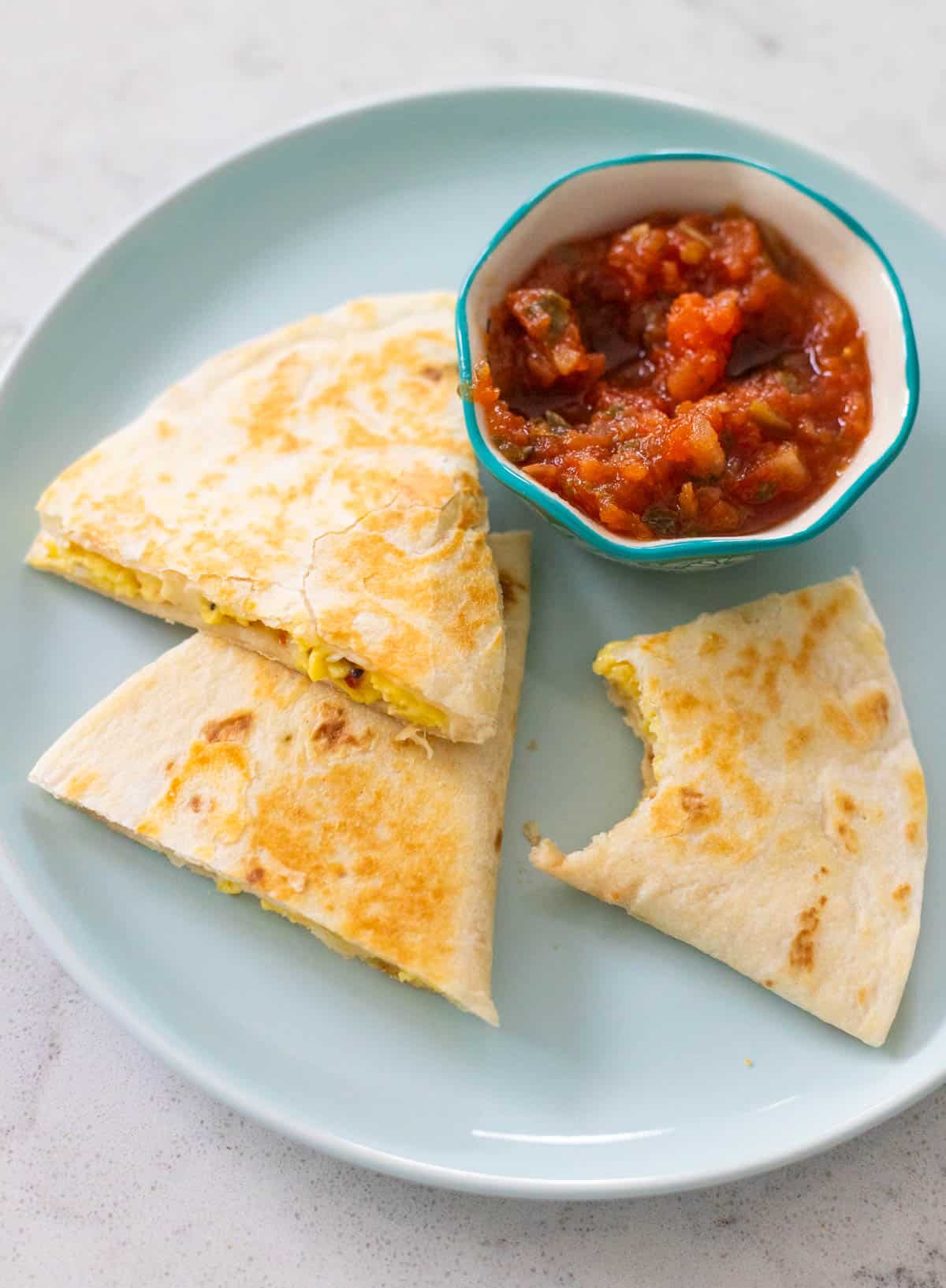 The cheese and egg quesadilla is being served on a plate with salsa. A bite is missing from one wedge.