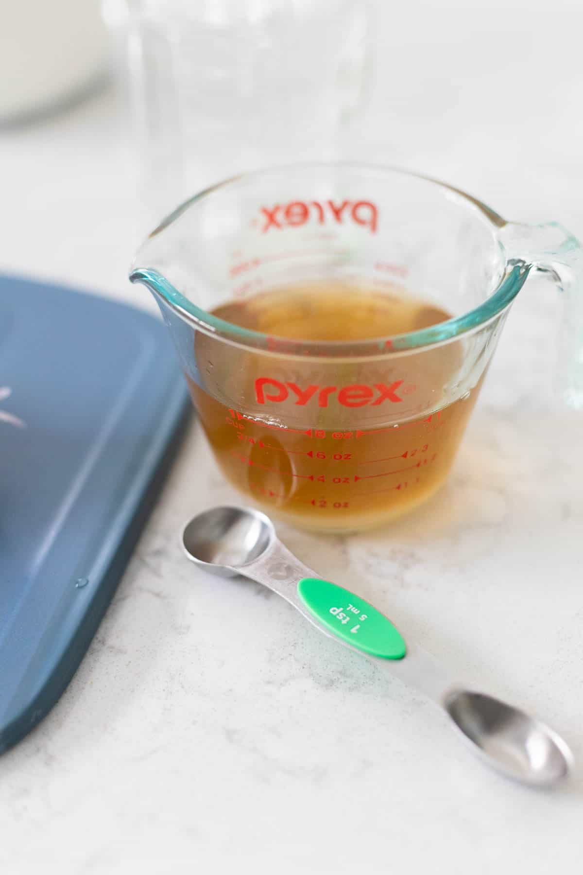 A measuring cup of vinegar has a measuring spoon on the side.