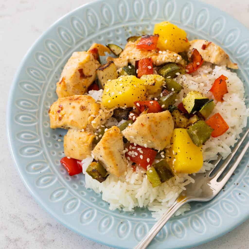 A blue plate has a bed of rice topped with chunks of chicken, pineapple, and roasted vegetables.