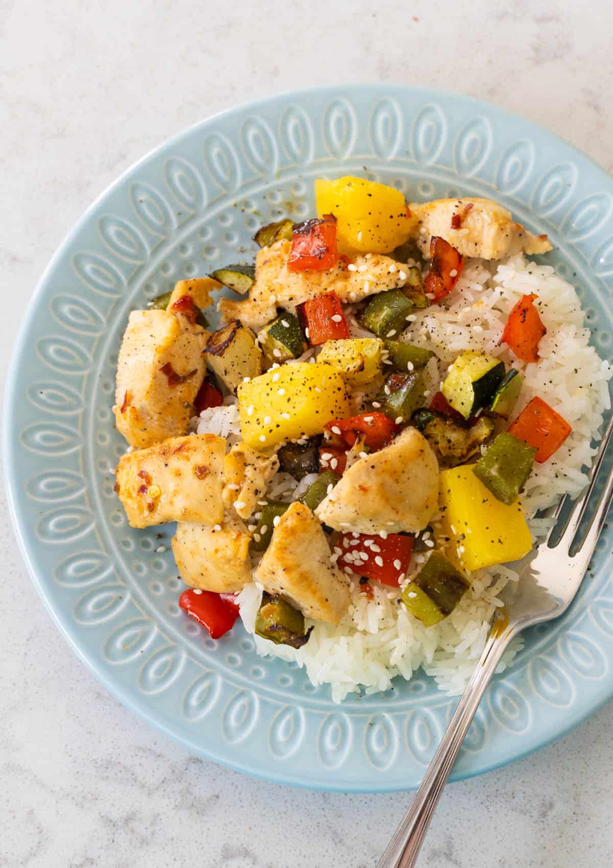 A blue plate has a bed of rice topped with chunks of chicken, pineapple, and roasted vegetables.