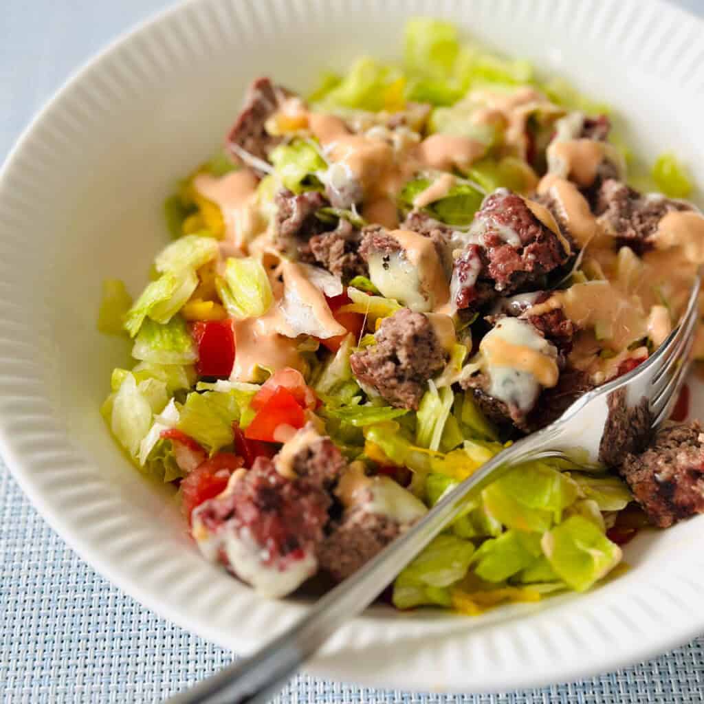A white bowl filled with lettuce and topped with a chopped up cheeseburger.