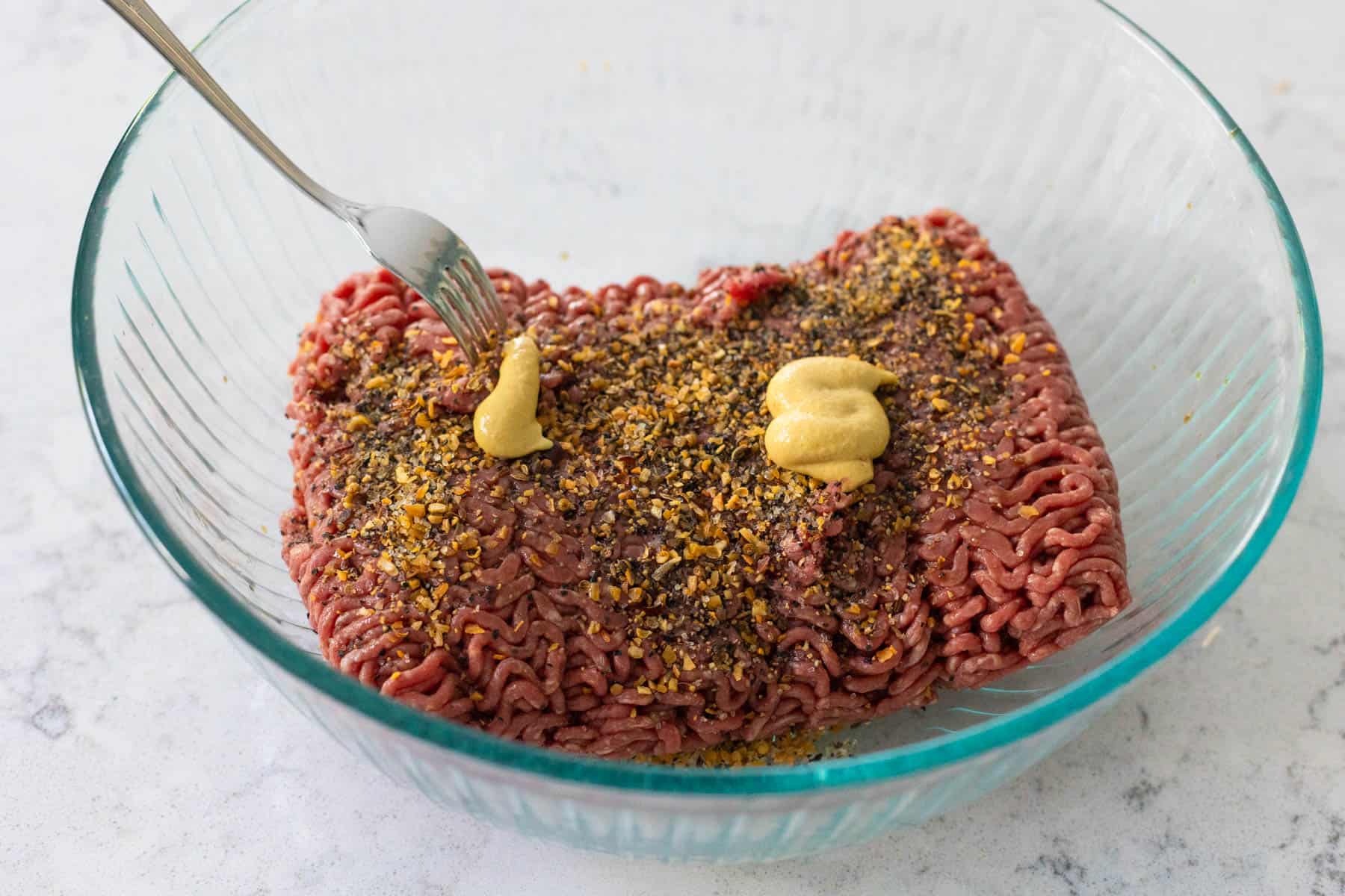 The ground beef is in a mixing bowl topped with seasonings and mustard.