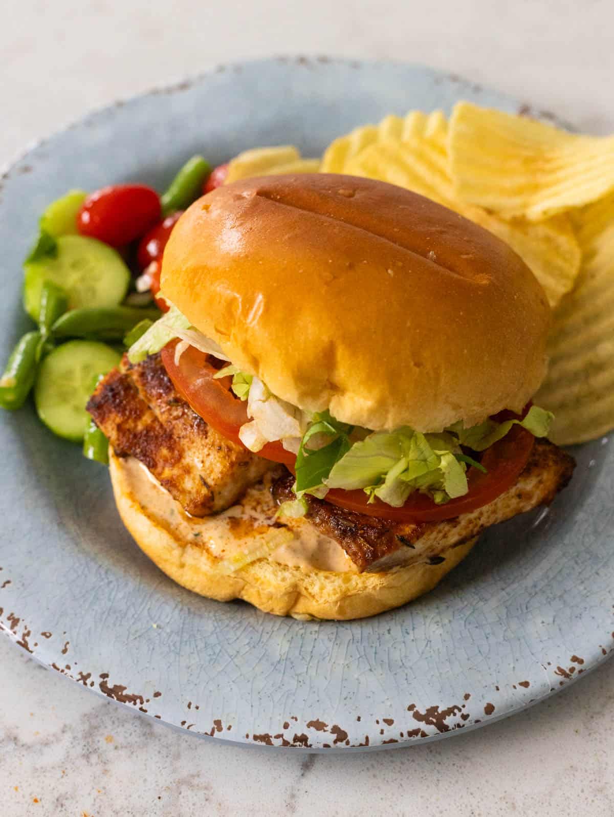 The fish sandwich is on a blue plate next to some potato chips and a raw veggie salad.