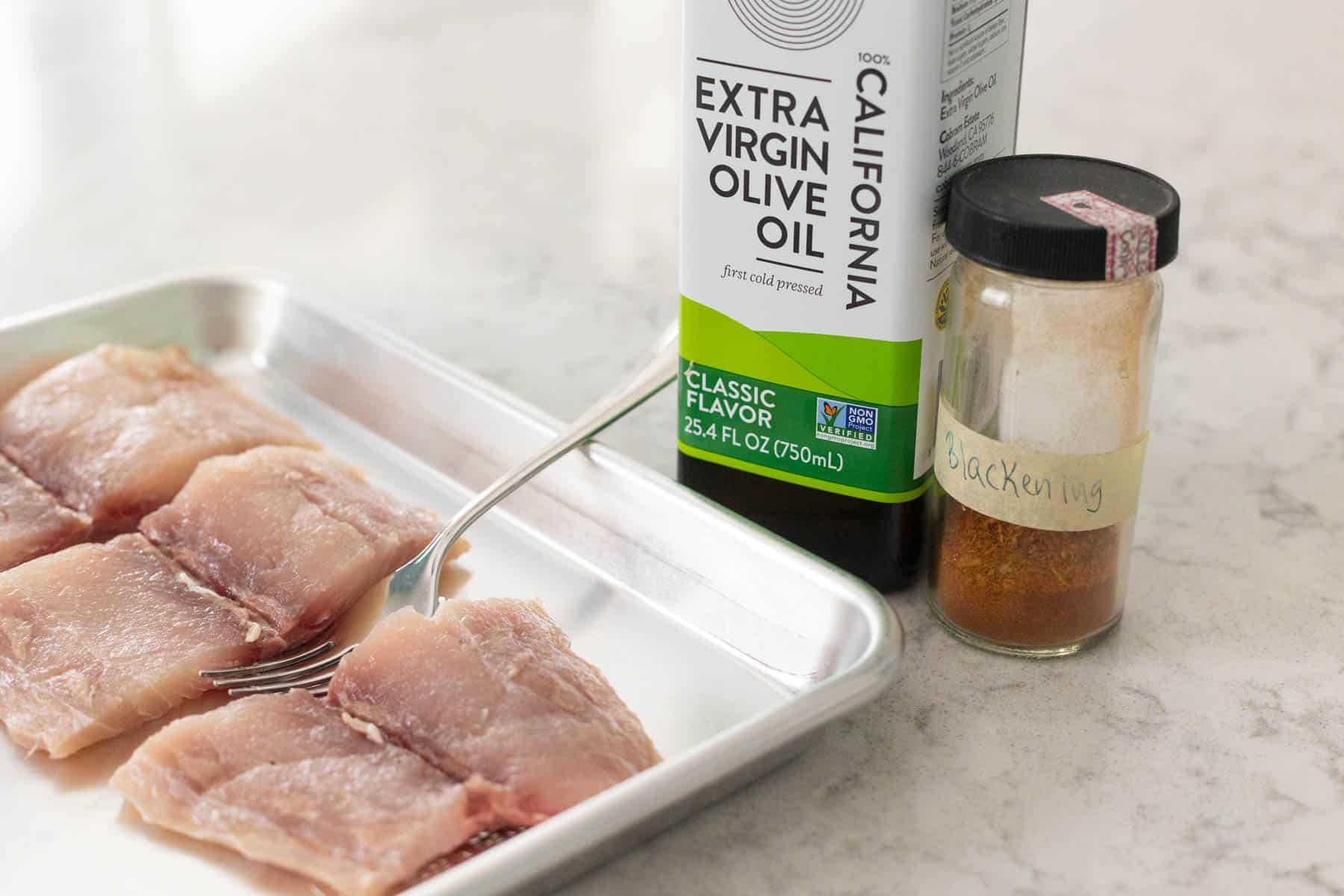 The raw fish is on a baking pan about to be seasoned. The olive oil and seasoning are on the side.
