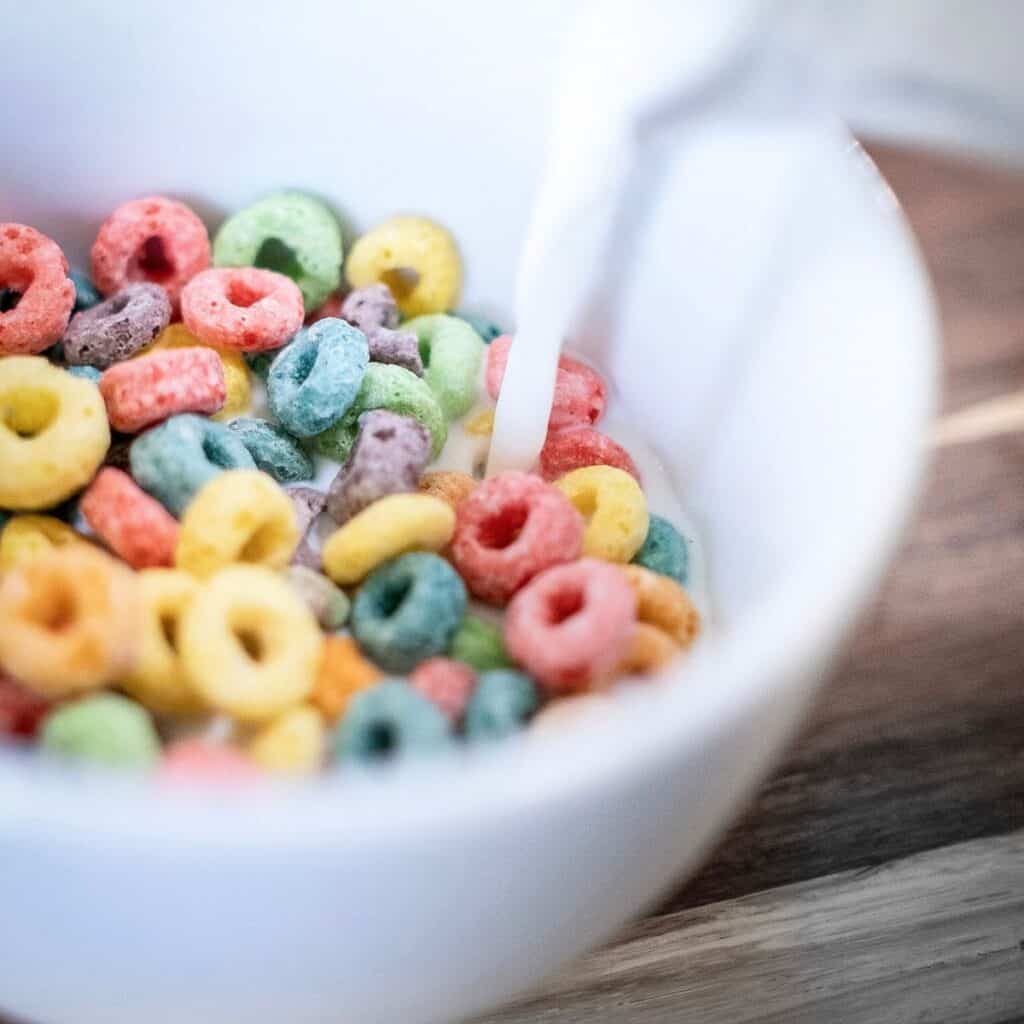 A white bowl filled with fruity cereal has milk being poured in.