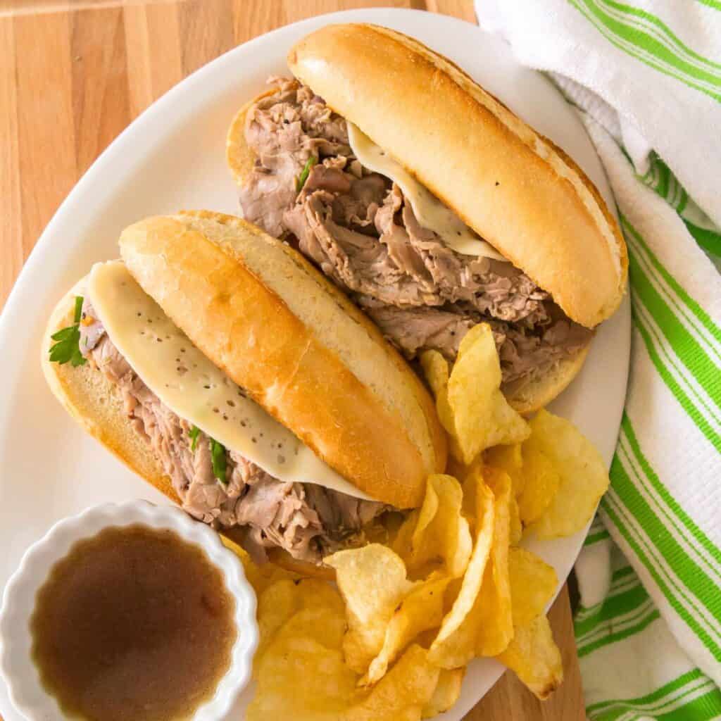 A french dip sandwich on a white plater next to a cup of beef broth. Potato chips are on the side.