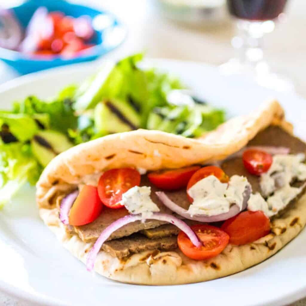 A gyro sandwich with fresh tomatoes and onions is on a pita next to a simple green salad.