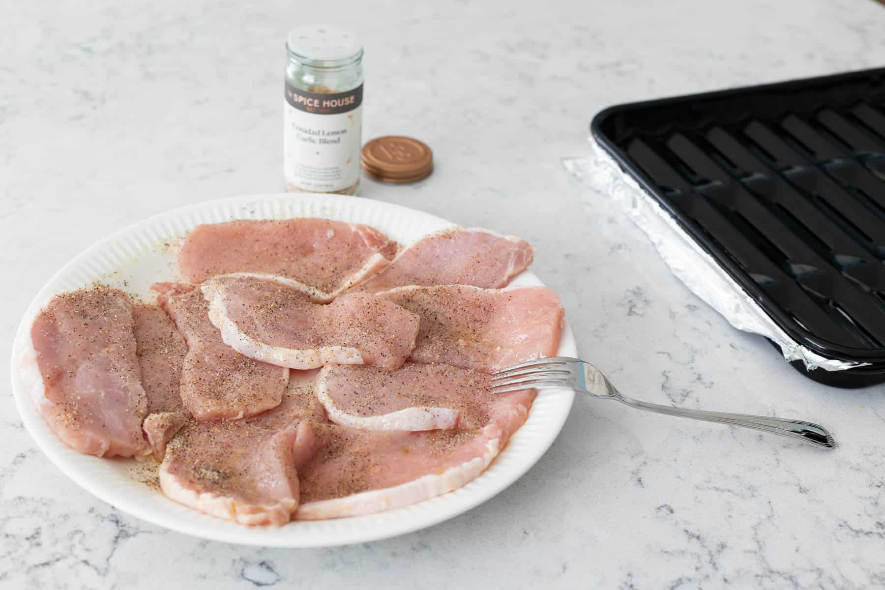 The pork chops are on a clean plate being sprinkled with seasoning.