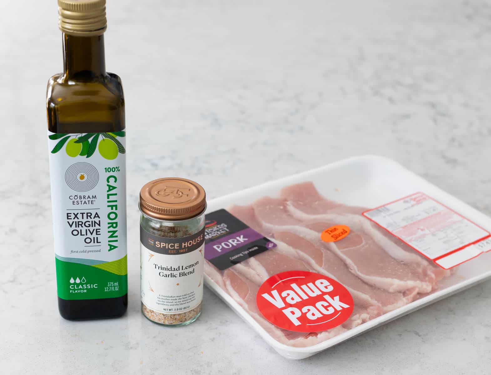 The bottle of olive oil, a jar of seasoning, and the package of pork chops are on the counter.