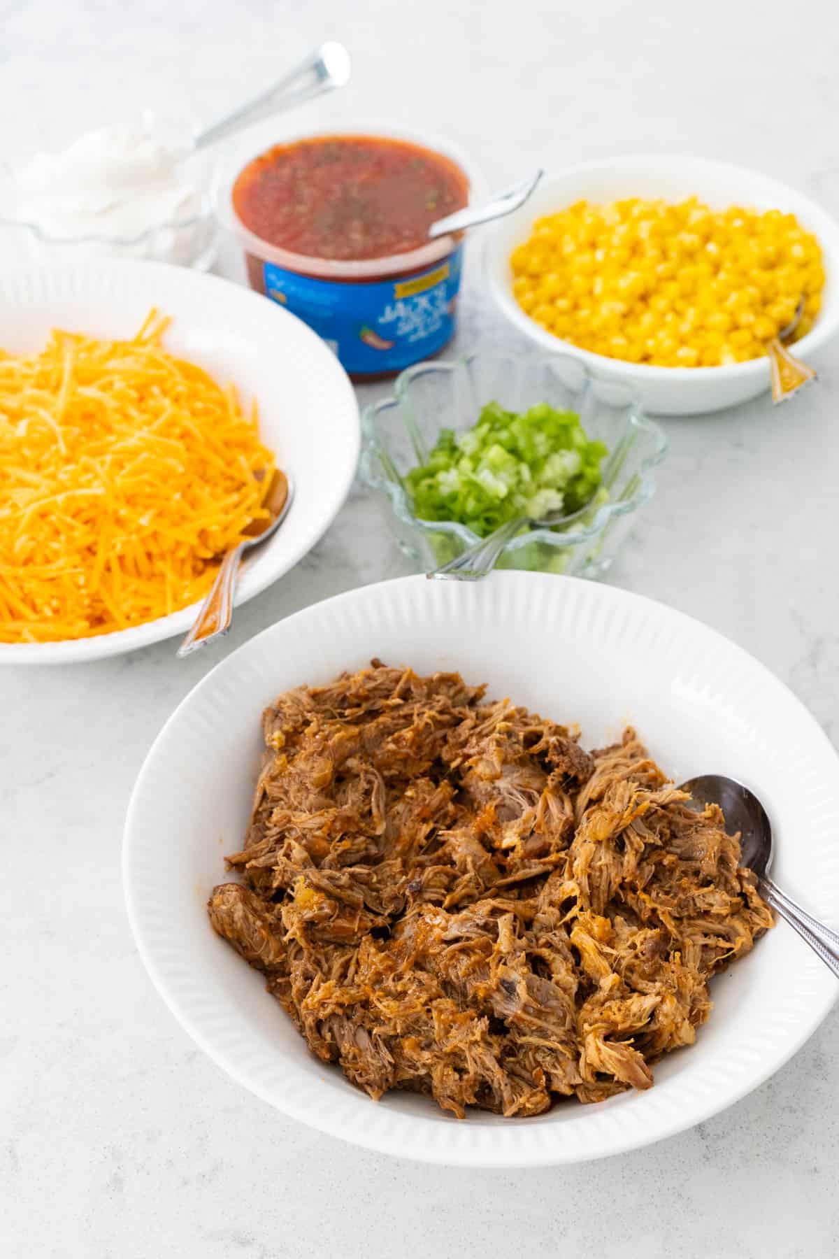 A bowl of shredded pork shoulder is in a white bowl surrounded by toppings to make nachos.
