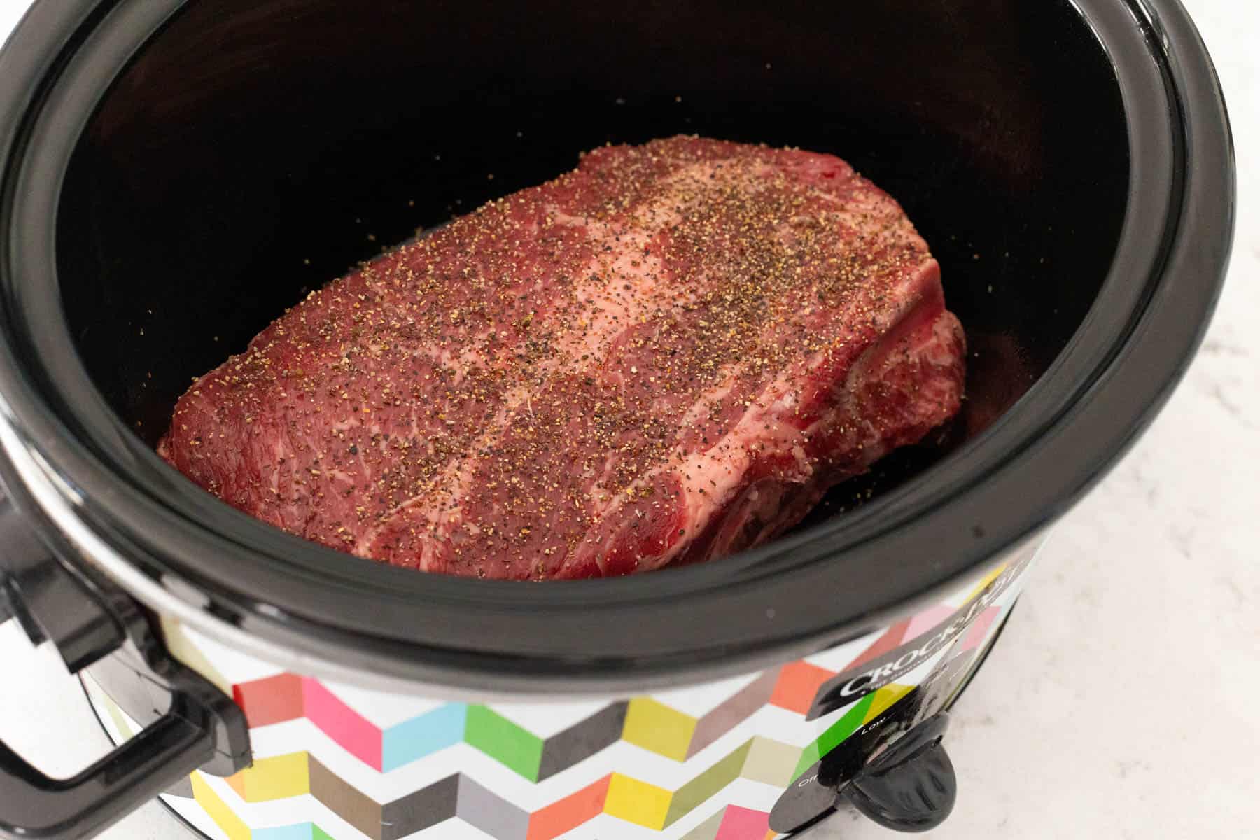 The beef roast is inside the slowcooker and has had seasoning sprinkled over the top.