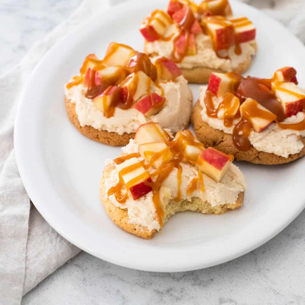 A white platter has cookies topped with frosting, chopped apples, and a drizzle of caramel.