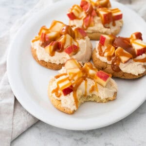 A white platter has cookies topped with frosting, chopped apples, and a drizzle of caramel.