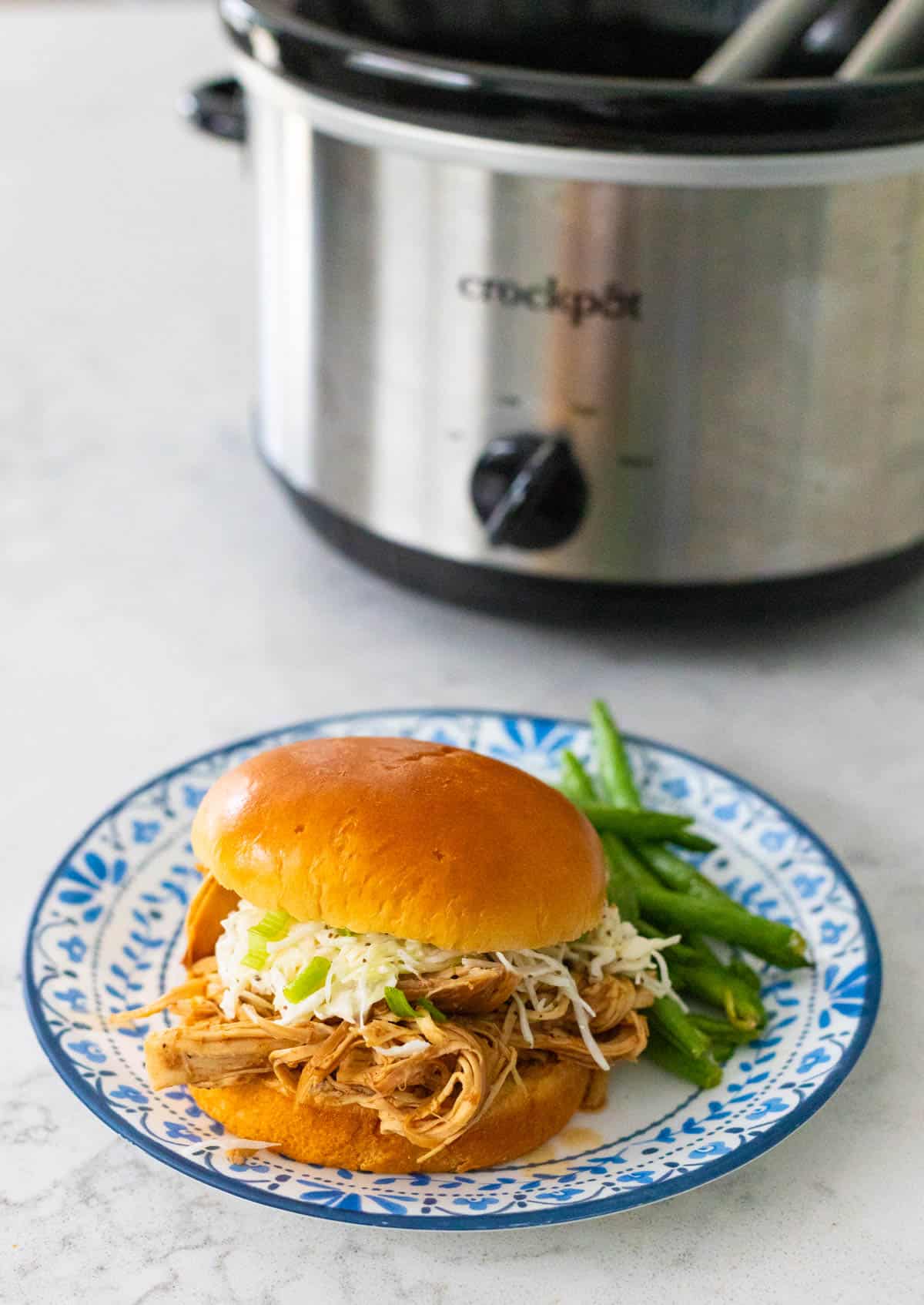 The shredded chicken sandwich has slaw and green onions on top, a serving of green beans on the side. The plate rests in front of the Crockpot where it cooked.