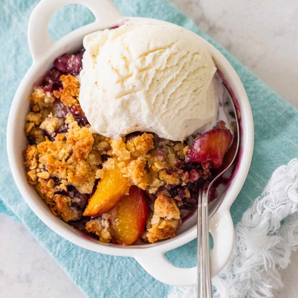 The white dish is filled with peach blueberry crisp and a scoop of vanilla ice cream.