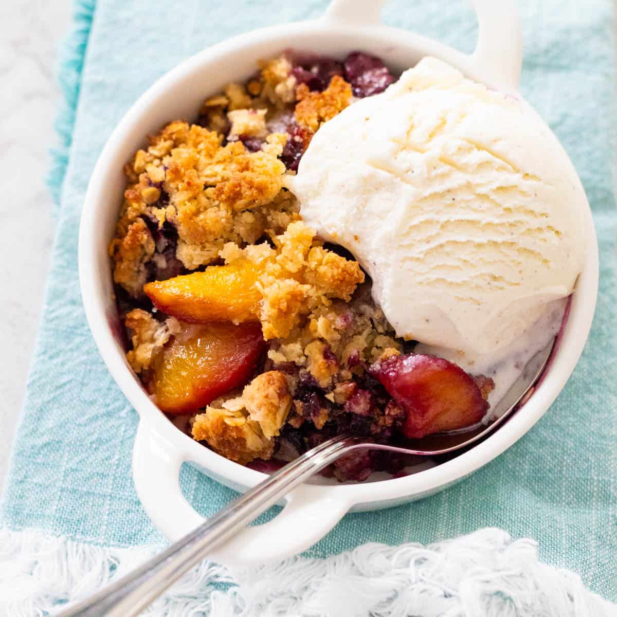 The white dish is filled with peach blueberry crisp and a scoop of vanilla ice cream.