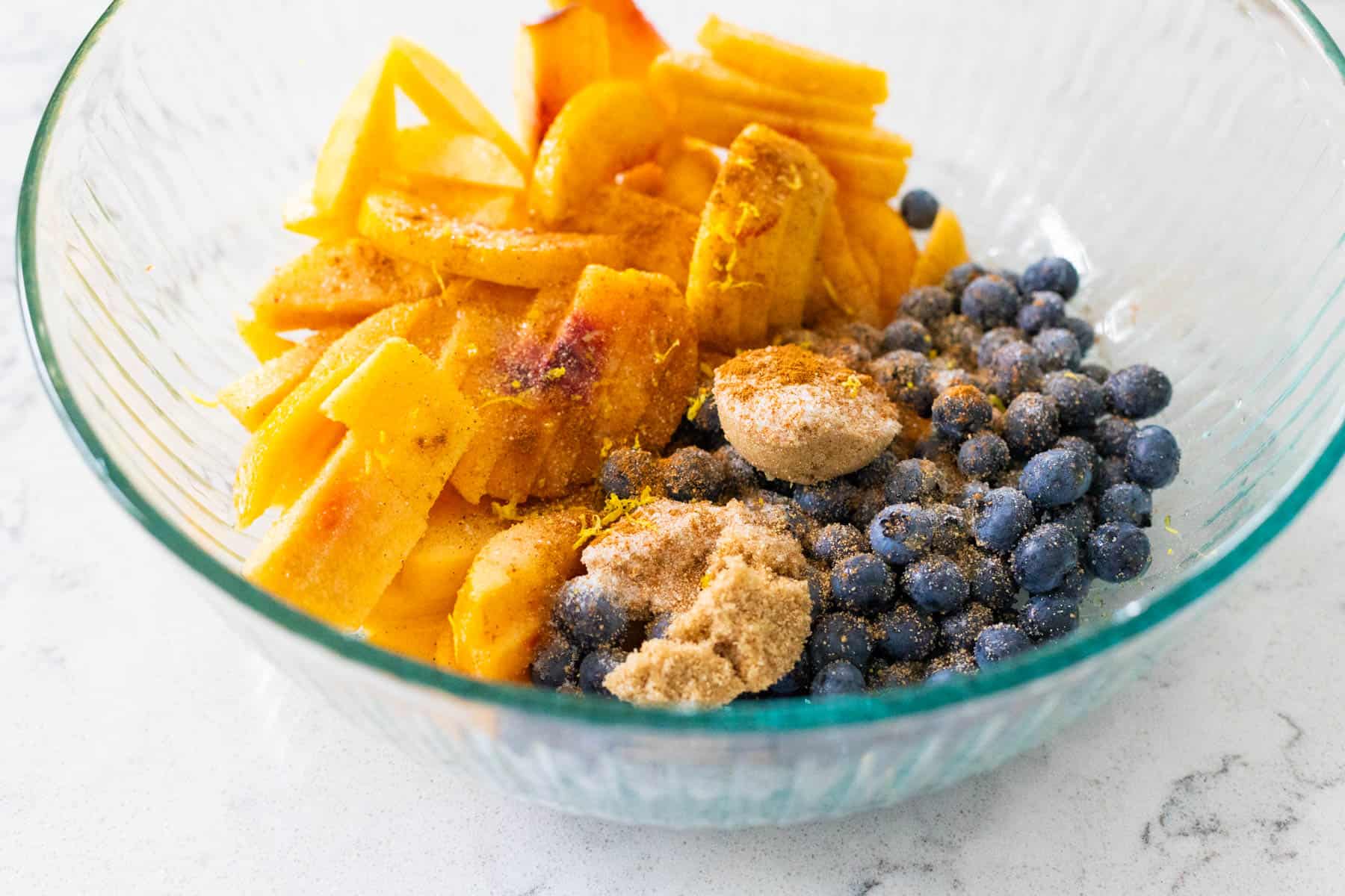 The peaches and blueberries are in a large mixing bowl with brown sugar and other seasonings sprinkled over the top.